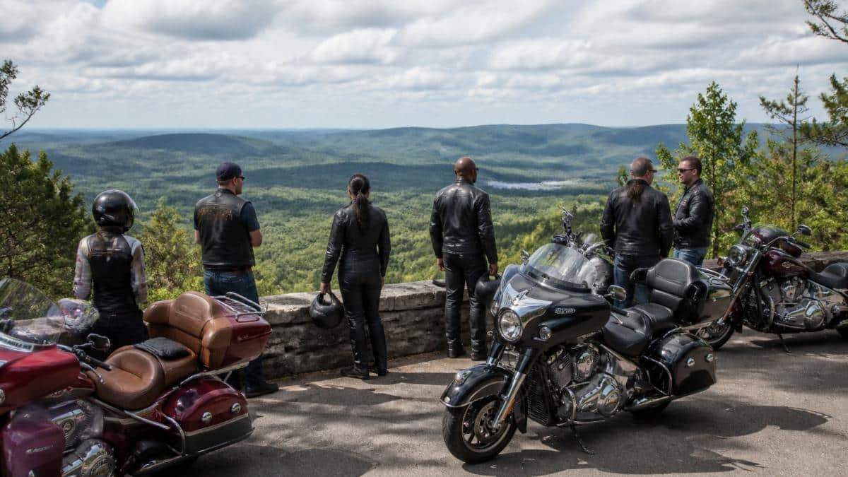 Group of Indian Motorcycle riders in leather gear gathered at a scenic overlook during an organized group ride, with forested landscape and distant mountains in background