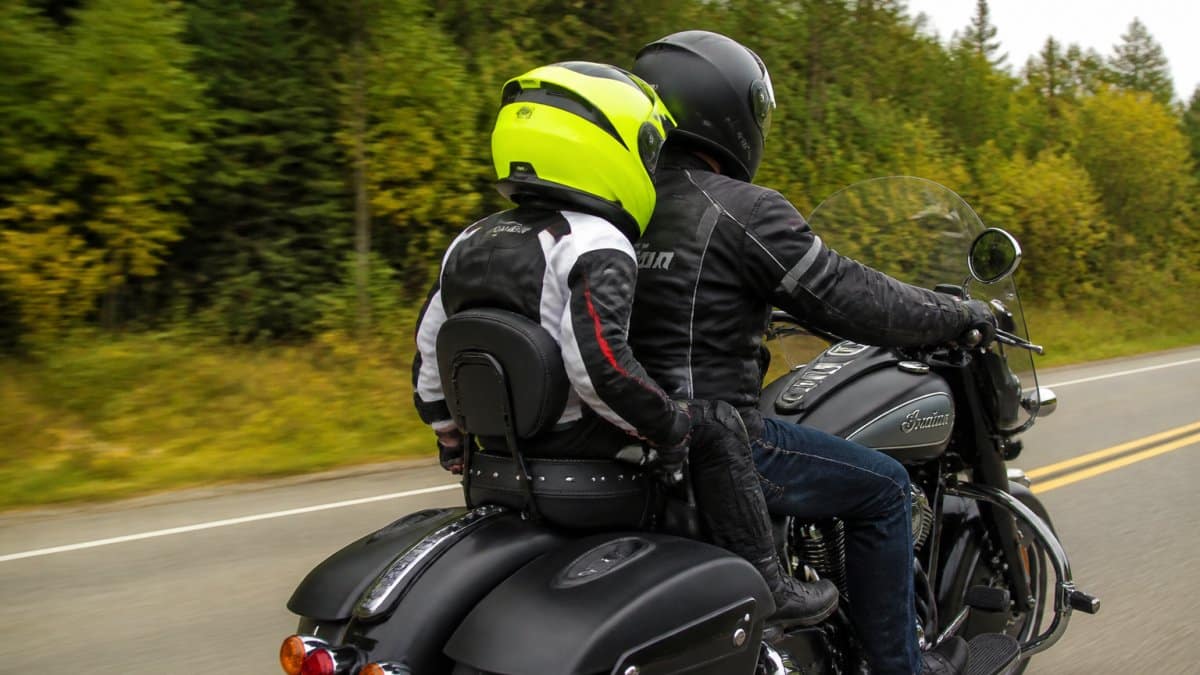 Child passenger on a motorcycle wearing a bright helmet and protective gear, seated on a passenger seat with both hands gripping grab handles, feet positioned on footrests, rider visible in front on open road with trees in background