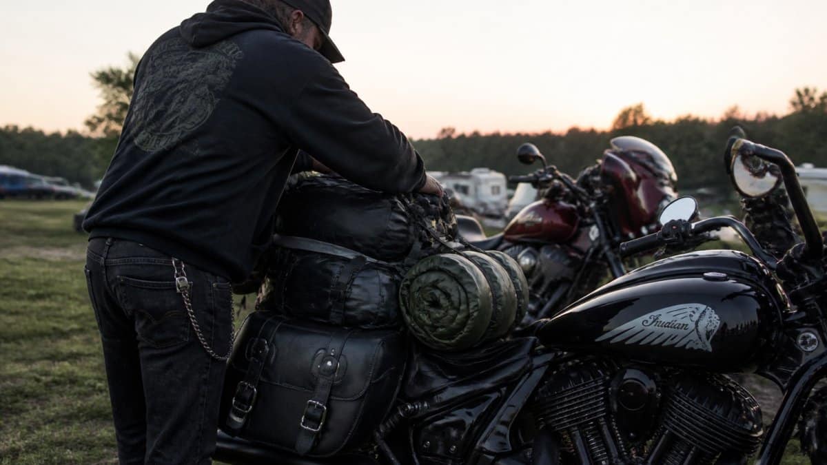 Rider loading camping gear onto a motorcycle at dawn, showing saddlebags, sleeping bag, and camping equipment organized and secured